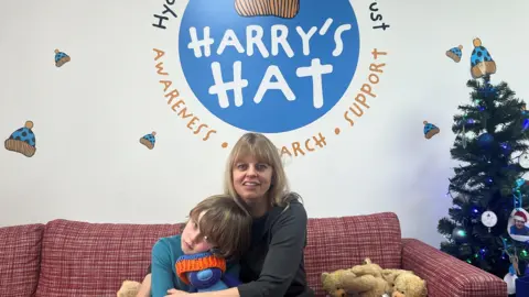 BBC/Julia Moore Woman with blonde hair hugging her son in a blue top, who's hugging a blue teddy bear in front of a logo that shows the charity's name, Harry's Hat.