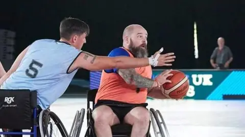 Dave Anderson Dave is in his wheelchair playing basketball. He's being tackled by an opposing player.