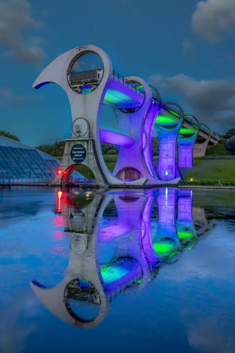 Dave Lynch The Falkirk Wheel illuminated in purple, green and red at night. The reflection is in the water and the sky is dark with clouds.