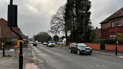 BBC Junction of Park Road and Coventry Road, Bedworth, Warwickshire. There are metal railings between the road and the pavement and tress on either side of the road on the bend of the junction. There are houses to the right of the image. 