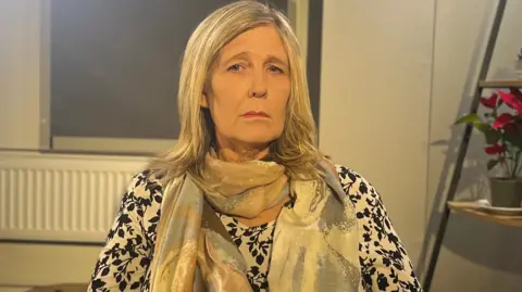 Vanessa is looking at the camera. She is wearing a black and white patterned long sleeve top and a brown, blue and cream scarf. She has shoulder length blonde hair. Behind her, there are shelves with plants on them and a window with blinds shut.