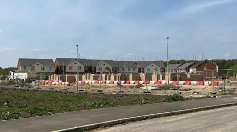 A building site in the foreground with piles of bricks behind small plastic barriers. Rows of new detached homes can be seen further back.