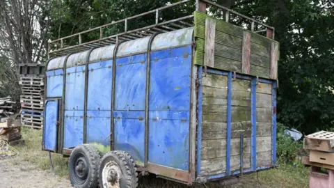 Crown Prosecution Service A large, enclosed trailer standing on a patch of dry grass and dirt in an outdoor yard surrounded by trees and miscellaneous stored materials.The trailer is painted predominantly blue and has a weathered appearance, with visible rust, dirt and wear on the metal frame. It is mounted on two axles with four large tyres, which are also worn and muddy. The structure is tall and box-shaped, with curved metal roofing panels running along the top. A metal rail framework extends above the roof, creating a raised rack-like structure around the upper edge of the trailer. The side of the trailer is made up of multiple blue metal panels secured by hinges and fasteners. Near the front left side, there is a small access door with a handle. The rear section consists of large wooden panels framed by blue metal supports. The wood is heavily weathered and discoloured, with patches of green staining or moss growth visible across the surface.