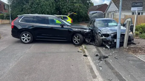 A car has rammed another car, leaving both badly damaged. The BMW has been shunted onto a pavement and has possibly hit a lamppost - its front and side are wrecked. The front of the unmarked police car is damaged. Another police car with markings is in the background.