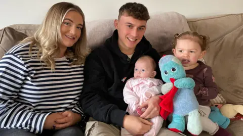 Lily has long blonde hair and is wearing a blue and white striped top. She is sitting on a beige sofa next to her partner Jacob who is wearing a black hooded top. He is holding their baby. Next to the baby is their older child holding an Iggle Piggle soft toy.