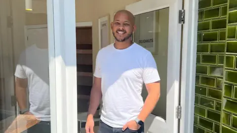 SAY Doughnuts Aaron, standing in a doorway of a shop, with green tiles on the walls outside. The door frame is white wood. He is wearing a white T-shirt, with jeans, a watch on his arm and is smiling and looking at the camera. He has a dark beard.