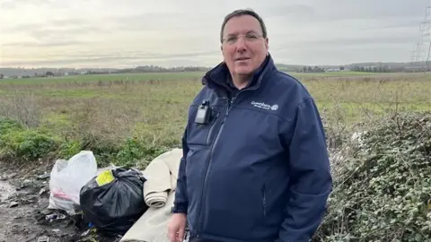 Tim Harris, Gravesham's Environmental Enforcement Manager, stands next to a flytipped carpet and black bin bags in fields overlooking Gravesend