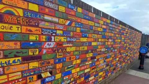 Somerset Council A colourful brick wall. Each brick is painted with pictures and messages in an array of colours - mostly yellows, blues, greens and reds.