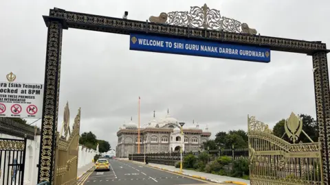 A gold and black arch over a road leading into the gurdwara, with a sign in white running on a blue background just below the arch saying "WELCOME TO SIRI GURU NANAK DARBAR GURDWAR"