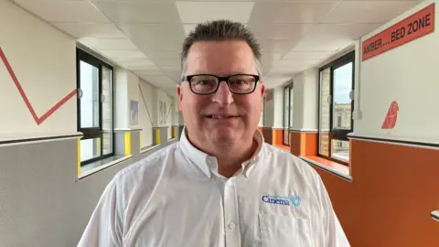 A man with short spikey hair and glasses wearing a white shirt with the words Together for Cinema stitched above the breast pocket. He is standing in a colourful hospital corridor.