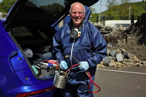 A man with white hair and glasses is wearing a blue body suit and blue plastic gloves, whilst holding a fumigating device