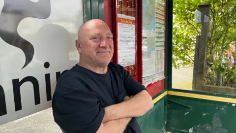 Peter sits with his arms folded, looking away from the camera. He is in a bus shelter, in the background there are bus tables and posters.