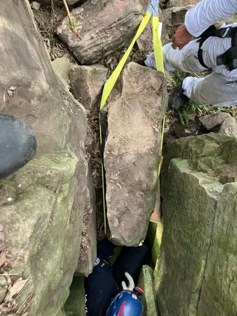 NSW Ambulance A rock with a yellow strap around it, lifted from between two other rocks. A person wearing a helmet can be seen in the crevice, while another person's legs can be seen next to the crevice