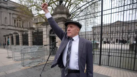 PA Media Michael Healy-Rae, a man with shaved head and a flat peaked cap, stands outside the tall iron gates of Dublin's Leinster House, with one fist raised above his head. He is wearing a grey/blue suit, a light shirt and a navy tartan tie. 