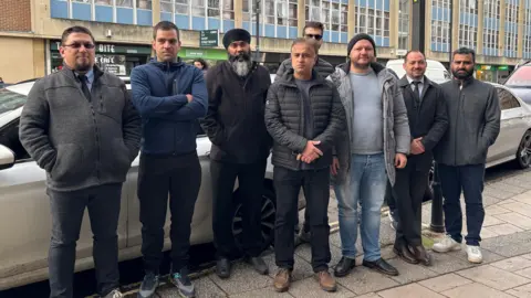 Eight Uber drivers standing in a line in front of their cars on a busy Bristol street. They are all men and are wearing a mix of thick fleeces and coats, and are looking towards the camera. Some of them are smiling slightly, as others look more serious. 