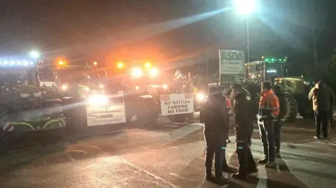 Philip Weston A row of tractors, some with banners saying "no British farming no food" line up in the dark outside the Asda depot. A number of farmers are standing around with their hands in their pockets.