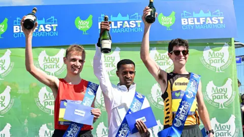 Pacemaker The three male winners of the Belfast Marathon hold up bottles of champagne in one hand and a blue envelope in the other. On the left is runner-up Aaron Hanlon, who is wearing a blue sash and a red athletic top and is smiling. In the middle is Tilahun Nigussie, who is wearing a white zip up jacket and is smiling at the camera. On the right is Stephen Connolly, wearing a yellow and black athlete best with 'Strident' written across the chest.