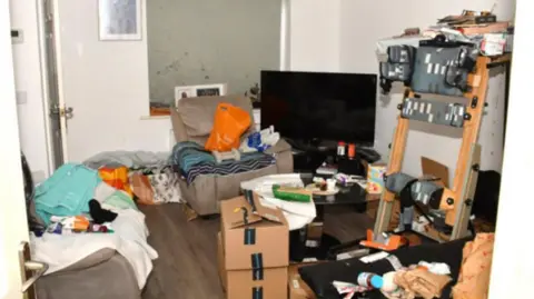 Merseyside Police A messy lounge area with a grey sofa and armchair, Amazon boxes on the floor and a coffee table strewn with various items. 