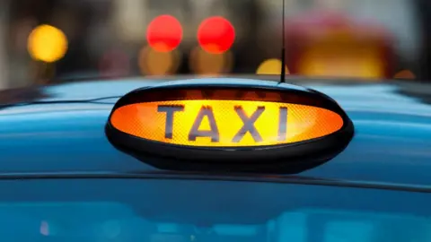 A stock image of the roof of a blue taxi with a "TAXI" light illuminated on it