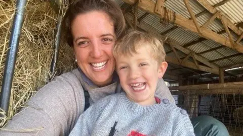 Handout A selfie taken by mum Lucy who is smiling next to her young son Ollie, who has short blonde hair. They are sitting next to a hay bale in a barn