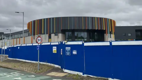 The top of the prison's entrance can be seen above blue, metal security fencing. The entrance has a curving wall with bands of bright colours.