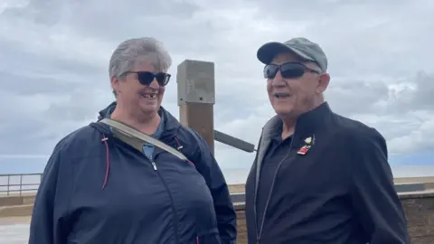 A woman with short grey hair and wearing sunglasses, looks towards a man with short grey hair wearing sunglasses. Behind them is the promenade and beach. They are smiling. Behind them is a cloudy grey sky.