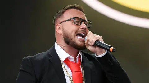 Getty Images Gareth tampil di Festival Leeds pada tahun 2022. Dia bernyanyi melalui mikrofon hitam, mengenakan blazer hitam, kemeja putih, dan dasi merah.