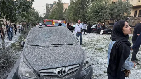 girl in headscarf stands to the right of a damaged car covered with dust