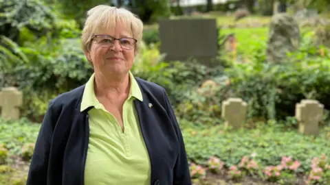 Helga has short, light-colored hair is standing outdoors in a garden or park area. She is wearing a light green collared shirt and a dark jacket. In the background, there are various plants, trees, and what appear to be gravestones or memorial markers.