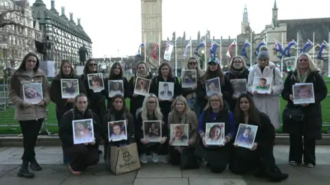 BBC A group of women pose together for a photograph. They are all holding a photograph of a child.