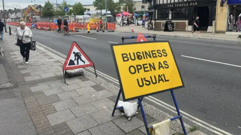 A yellow road sign says "Business open as usual" and it sits just in front of a roadworks sign, both are next to the road with the roadworks visible in the distance and The Temple Bar just next to the works.