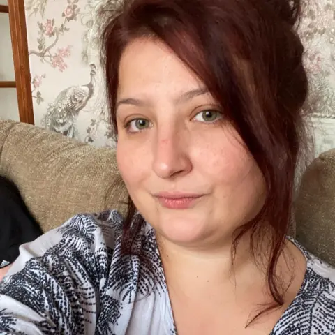 Supplied Jamie, a woman with auburn hair wearing a black and white top, she is sitting on a beige sofa with patterned wallpaper seen behind her 