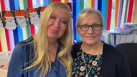 A woman with blond hair and a blue top stood beside an older lady with grey hair and black framed glasses in a dark top with flowers on. Both are stood in front of a multi coloured striped wall