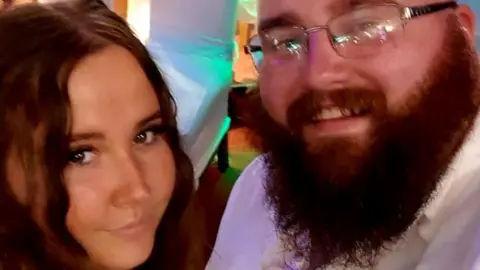 Nicole and Darren smile at the camera, she has long brown hair. He has a long brown beard, wears glasses and a white shirt