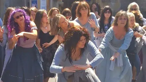 Cheryl Sprinkler A group of women dressed in blue dresses is dancing in the sun.