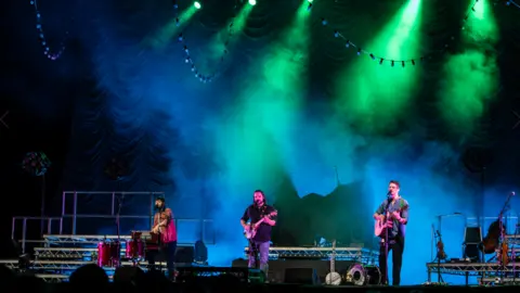 Bristol Harbour Festival Image of a three man band playing on a wide stage. The lighting is blue and green behind them.