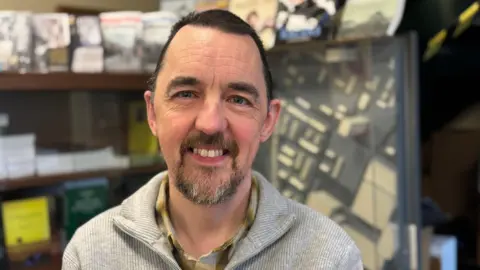 A man sits in an office. He is smiling and has dark hair and a greying beard. He's wearing a grey jumper over a yellow toned shirt.behind him and slightly out of focus is a shelf of books and pamphlets and a framed model of a road system. 