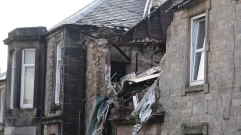 PA Media A grey sandstone building with a large hole on the upper floor and the roof. Debris from inside the flat can be seen sticking out of the gap.