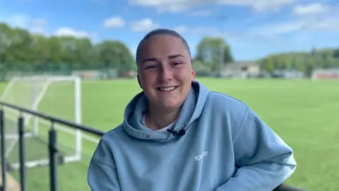 Chloe is standing on the side lines of a football pitch wearing a blue hoodie, her hair is tied back and she is smiling at the camera. There is a football goal in the background.