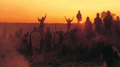 Mensch Films/Robin Mahoney An image at sunrise with lots of people standing looking towards the horizon on a field in semi-darkness. There is an orange glow to the sky and two people are silhouetted in the distance with their arms in the air.