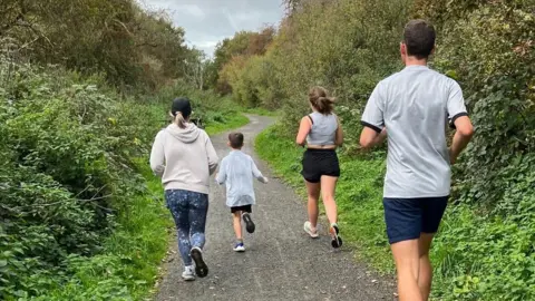 Family photo Four people are jogging along a wide dirt path surrounded by dense greenery on both sides. The path curves slightly ahead and is lined with trees and bushes. The sky is overcast with grey clouds. The runners are dressed in casual athletic wear: one in a hoodie and patterned leggings, another in a light top and shorts, and two others in shorts and T-shirts. They are moving away from the camera toward the bend in the trail.