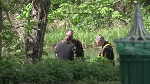 BBC Three police divers beside a river. They are beside trees and foliage and a green bin is in the front of the picture. 