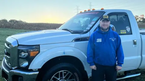 Ben Maurer Ben Maurer wears a blue jacket and a black CAT baseball cap in front of a white pickup truck