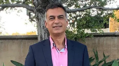Dr Sanjiv Nichani Dr Nichani is wearing a pink shirt with a navy blue suit jacket. He has dark brown hair which is greying at the sides.