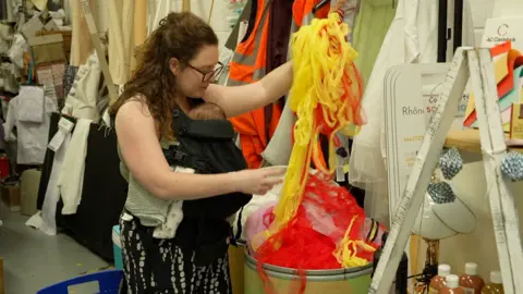 A woman with a very young baby in a baby carrier inspects brightly coloured nylon material in a round storage bin.