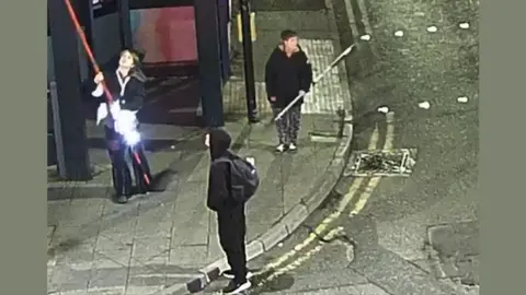 A CCTV image shows three people standing on a well-lit street corner at night. One person is holding a long red pole, another is holding a slightly shorter grey-coloured pole. A third person, at the forefront of the image, wears a dark top with the hood up, and is carrying a black backpack.
