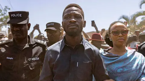 Bobi Wine in a black shirt flanked by a police officer and his wife in a blue shawl on voting day.