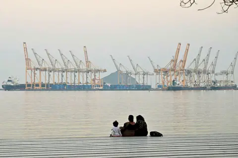 AFP via Getty Images A family sits against the backdrop of a dockyard off the coast city of Fujairah, in the Strait of Hormuz in the northern Emirate on February 25, 2026. 