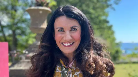 A woman with dark brown hair smiles at the camera, wearing a floral dress.