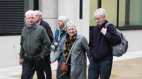 Eddie Mitchell A group of three men and two women, all with grey hair, walk together outside a building. 